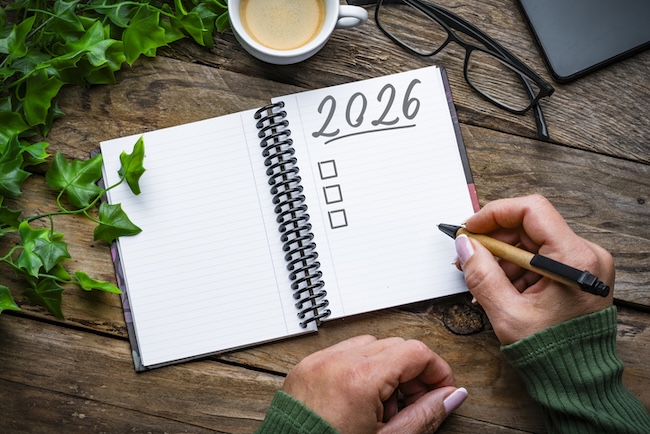 Aufgeschlagener Block mit Überschrift "2026". Person setzt mit einem Kugelschreiber zum Schreiben an.