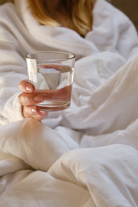 Frau sitzt mit einem Glas Wasser im Bett und streckt es dir entgegen