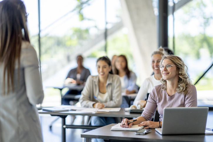 Eine Gruppe Erwachsener sitzen an Tischen und schreiben etwa auf einen Block. Ihre Laptops stehen offen auf dem Tisch