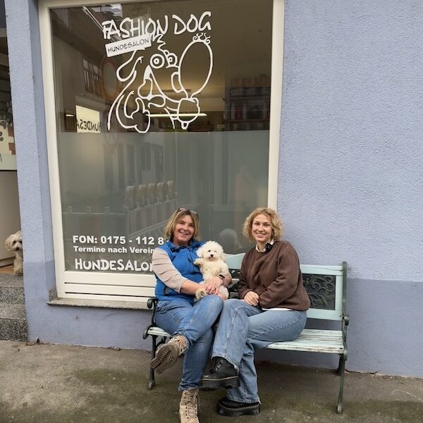 Lina und Claudia sitzen auf einer weißen Bank und Claudia hält ihren kleinen Hund auf dem Arm. Im Hintergrund sieht man das Logo vom Hundesalon Fashiondog.