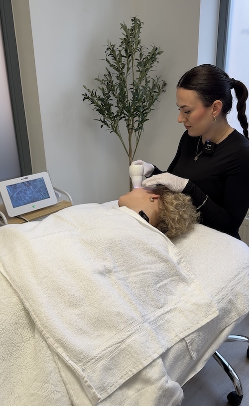 Lina liegt auf einer Behandlungsliege und hat den Kopf zur Seite gelegt. Sie schaut in Richtung eines Desktops. Auf dem Desktop sieht man die Haut von Lina vergrößert. Luana sitzt gebeugt hinter der Liege und hält das Vergrößerungsgerät an Lina's Gesicht.