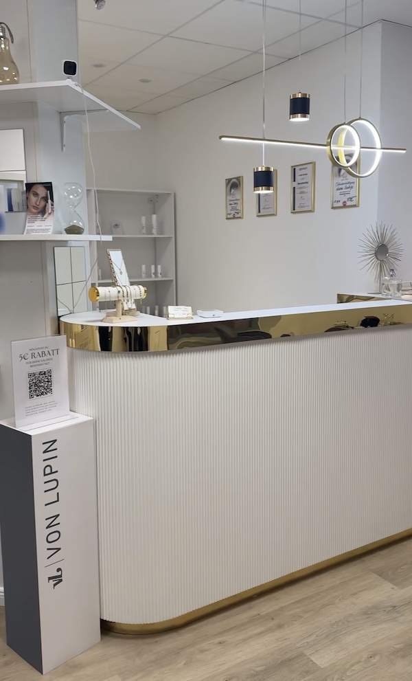 Main Beauty's weiß-goldenfarbige Rezeption mit Schrank voller Pflegeprodukte im Hintergrund.