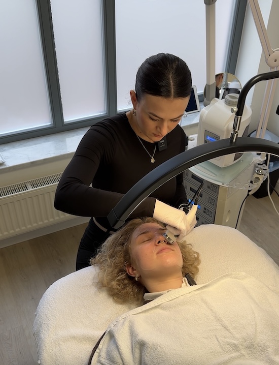 Lina liegt auf einer Behandlungsliege. Luana sitzt gebeugt hinter ihr und hält das Ultraschallgerät an Lina's Gesicht.