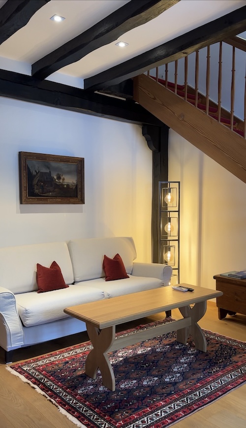 Weiße Couch mit Holztisch auf rotem Teppich. An der Seite eine Holztreppe.