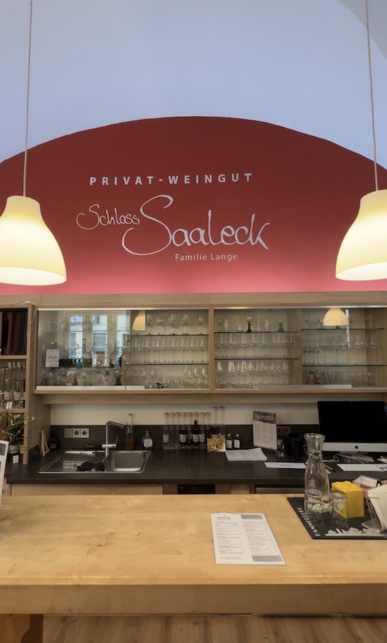 Die Holztheke vom Weingut Lange mit Gläsern im Hintergrund. An der Wand im Hintergrund steht "Privat Weingut, Schloss Saaleck, Familie Lange"
