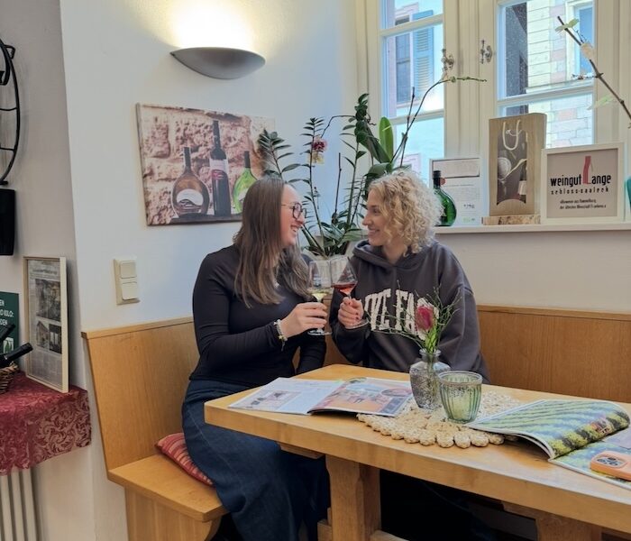 Lina und Hanna sitzen in einer Sitzecke und stoßen lächelnd mit ihren Weingläsern an.